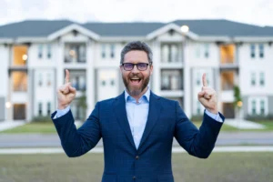 A cheerful real estate agent in a blue blazer stands in front of a modern apartment complex, smiling and pointing upward, expressing enthusiasm and success in the real estate industry.